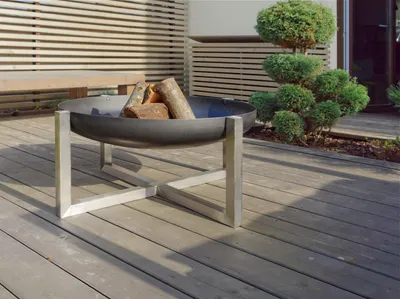 A fire pit with logs on a wooden deck, with a decorative bush and wooden fence in the background.