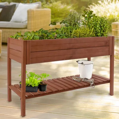 A wooden raised garden bed filled with various plants, placed on a wooden deck with outdoor furniture in the background.