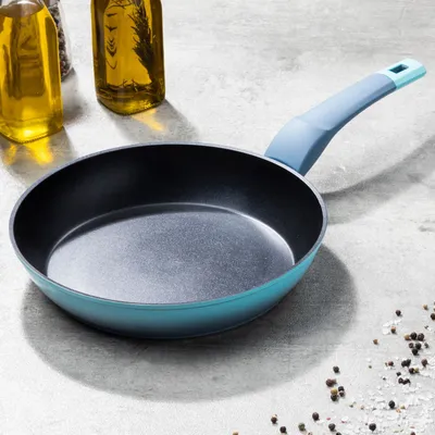 A turquoise frying pan with a light blue handle sits on a grey surface, with bottles of oil and peppercorns in the background.