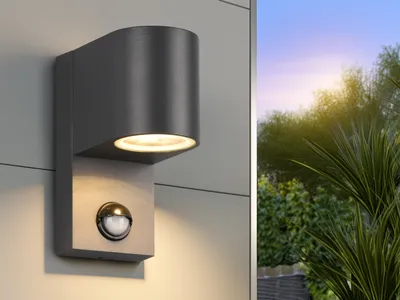 A modern gray outdoor wall light with a motion sensor is mounted on a light gray wall. The background shows a lush green garden with trees and a sunset sky.