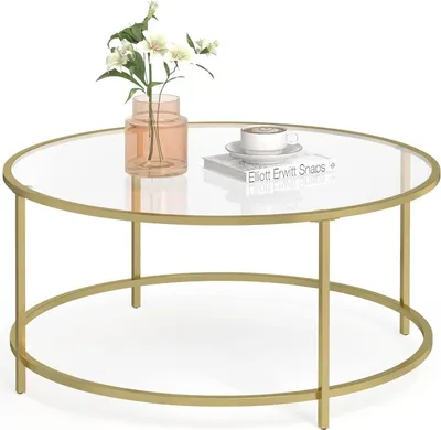 A round glass-top coffee table with a gold metal frame, a vase with flowers, and a book with a cup of coffee on it.