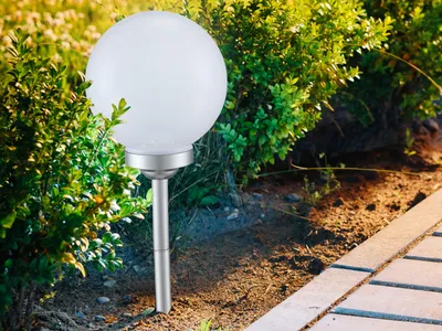 A solar garden light with a white globe sits among green bushes next to a paved walkway.