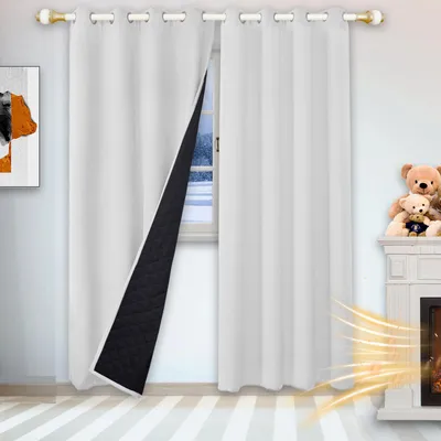 A white thermal curtain is partially open, revealing its black quilted lining. The curtain is hung on a rod with gold eyelets. A window with a winter scene is visible behind the curtain. A fireplace with teddy bears on top is on the right side of the image