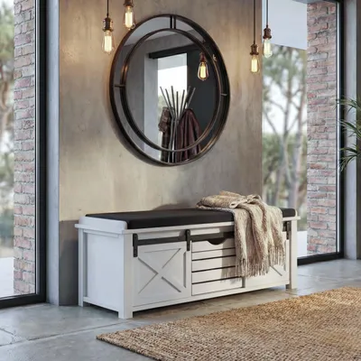 A white storage bench with a black cushion and a knitted throw is placed in an entryway with a large round mirror above it.