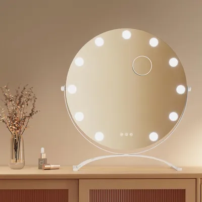 A round makeup mirror with LED lights sits on a wooden dresser, next to a vase of dried flowers and some cosmetic products.