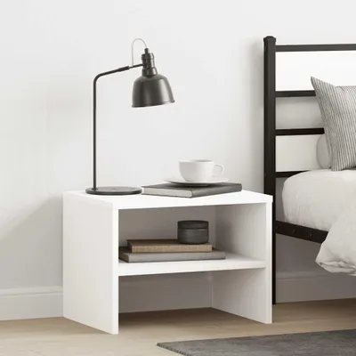 A white bedside table with a lamp, books, and a cup on it, next to a bed.
