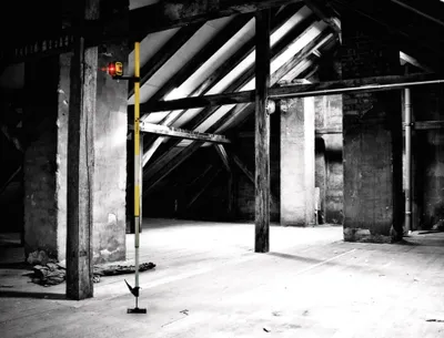 Interior of an old attic or unfinished room with wooden beams and concrete walls, featuring a yellow telescopic pole with a red light on top.