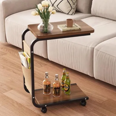 A C-shaped side table with a walnut finish is placed next to a sofa, holding a vase of flowers, a book, and a mug on its top surface. The lower shelf holds four bottles of beverages. The table has a fabric magazine holder on its side and wheels at the base