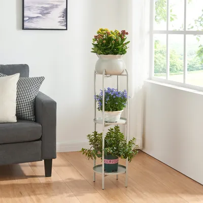 A plant stand with three potted plants is placed next to a gray sofa in a living room, with a window in the background.