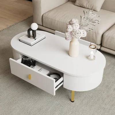 A white oval coffee table with gold accents, featuring an open drawer with a camera and headphones inside, is placed in a living room setting next to a sofa.