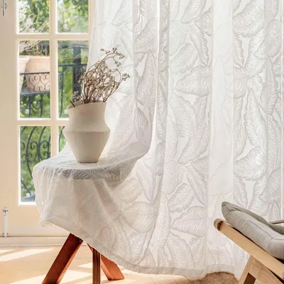 A sheer white curtain with a leaf pattern hangs in front of a window, with a small table holding a vase of dried flowers.