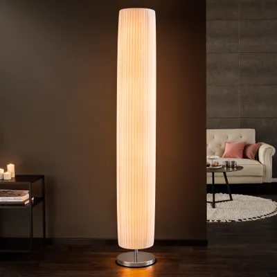 A tall, white, pleated floor lamp stands in a living room setting, next to a dark side table with candles. In the background, a sofa and coffee table are visible.