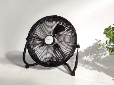 A black floor fan is shown in a studio setting with a white background and some shadows. A green plant is visible on the right side of the image.
