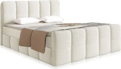 A modern, cream-colored boxspring bed with a bouclé fabric finish, shown from a side-front angle with two pillows and a throw blanket.