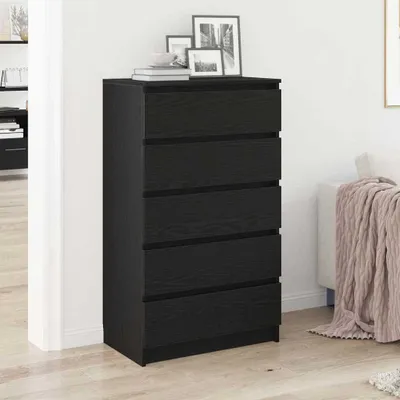 A black chest of drawers with five drawers, standing in a room next to a white wall and a couch with a blanket.