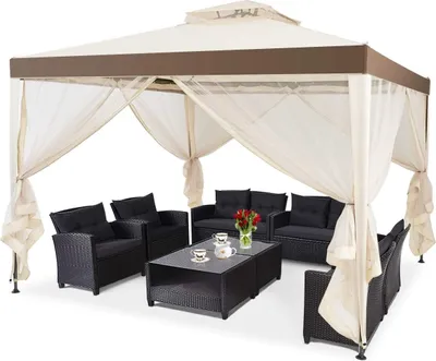 A beige and brown gazebo with sheer curtains, set up with black outdoor furniture and a coffee table with a vase of flowers and teacups.