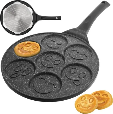 A close-up view of a non-stick pancake pan with seven smiley face molds, one of which contains a cooked pancake. Two additional smiley face pancakes are shown in the foreground.