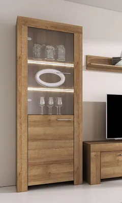 A tall, narrow glass-fronted cabinet with wooden doors and shelves, illuminated by LED lights, stands in a room next to a television stand.