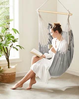 A woman is relaxing in a hanging chair, reading a book and holding a mug.