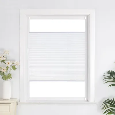 A white pleated blind is installed in a white window frame. A vase with flowers is on the left, and a plant is on the right.