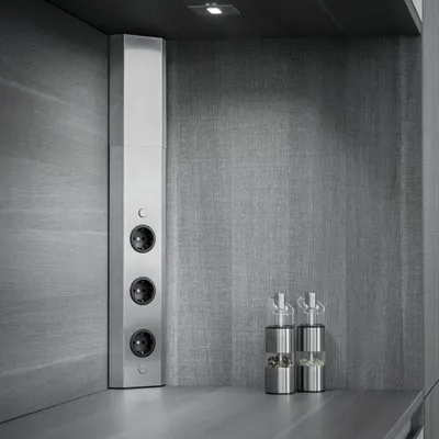 A close-up shot of a silver corner power outlet strip with three black sockets, mounted on a textured gray wall in a kitchen or modern interior setting. Two stainless steel and glass salt and pepper grinders are visible on the countertop in the foreground.