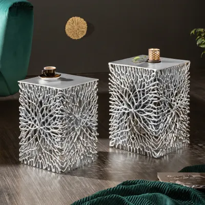Two silver side tables with intricate, branch-like bases are placed on a wooden floor next to a green armchair. One table has a coffee cup on it, and the other has a candle.