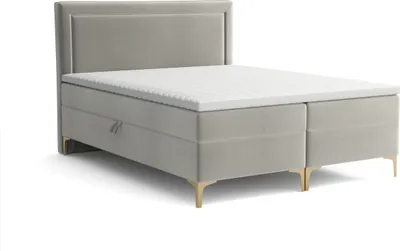 A studio shot of a light gray upholstered box spring bed with a white topper and gold legs, against a white background.