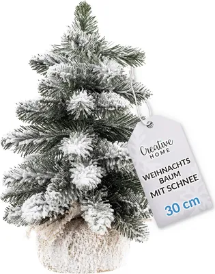 A small, artificial Christmas tree with snow-like flocking, placed in a pot and photographed against a white background. A tag is attached to the tree.