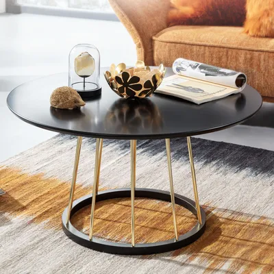 A round black coffee table with gold legs is placed on a rug in a living room setting, with a sofa in the background.