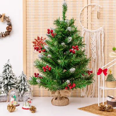 A small, decorated artificial Christmas tree is displayed on a white surface with other festive decorations in the background.