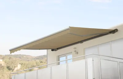 A beige retractable awning is extended over a balcony, with a window and railing visible below. The background shows a slightly blurred landscape and sky.