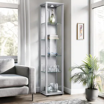 A tall, narrow glass display cabinet is shown in a living room setting, next to a sofa and a large window. The cabinet contains several decorative items.