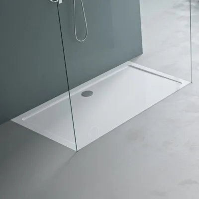 A modern white rectangular shower tray with a drain, enclosed by glass panels and set against a grey wall and concrete floor.