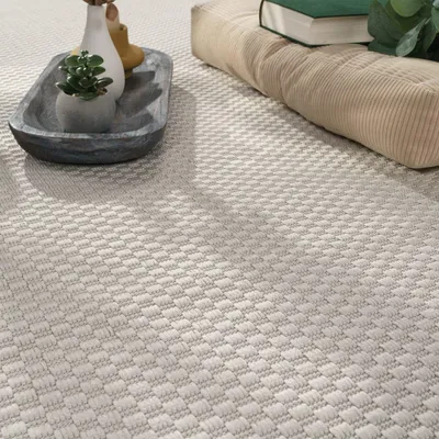 A close-up view of a cream-colored rug with a subtle geometric pattern, featuring decorative items like vases and a small plant on a tray in the foreground, and a cushion with books in the background.