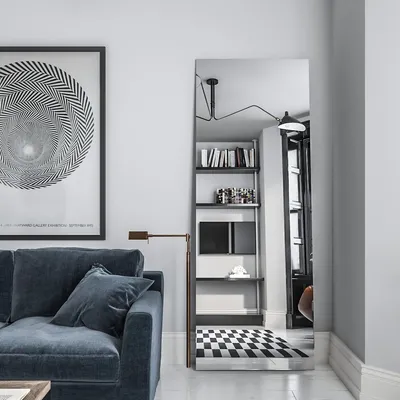 A modern living room with a blue sofa, a large framed artwork, and a tall floor mirror reflecting a bookshelf and a checkered rug.