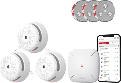 A product shot of a smart smoke detector bundle, including three smoke detectors, a base station, a smartphone displaying an app interface, and mounting accessories, all arranged on a white background.