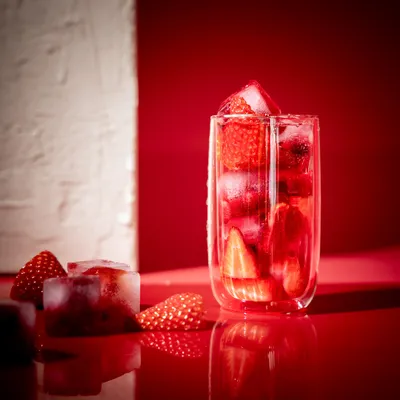 A glass of strawberry-infused water with ice cubes and strawberries on a red surface.