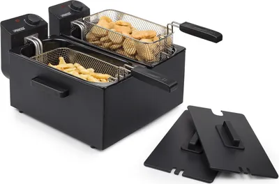 A black double deep fryer with two baskets, one filled with french fries and the other with chicken nuggets. Two black lids are placed in front of the fryer.