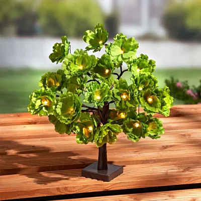 A decorative LED tree with green flowers and yellow lights sits on a wooden surface.
