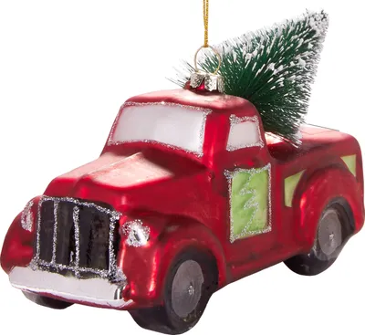 A close-up studio shot of a red glass Christmas ornament shaped like a pickup truck with a small green Christmas tree in the bed.