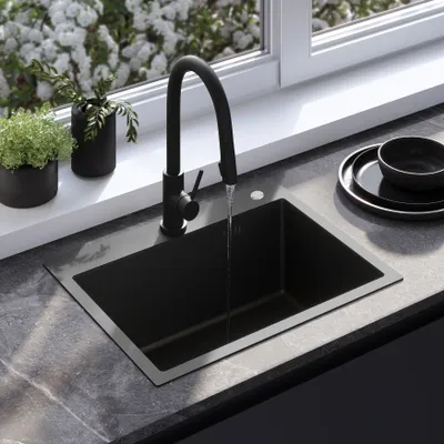 A black kitchen sink with a black faucet is shown with water running into it. There are plants on the windowsill behind the sink and a stack of black plates to the right.