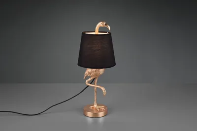 A gold flamingo-shaped table lamp with a black lampshade sits on a gray surface against a dark gray background.