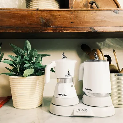 A white Ariete espresso maker and milk frother/water kettle set on a marble countertop, with a potted plant to the left and wooden shelves above.