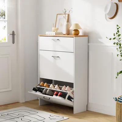 A white shoe cabinet with wooden top and two drawers, placed in a hallway next to a door and a wall with decorative molding.