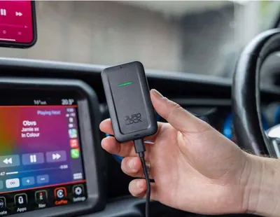 A hand holding a black Quad Lock wireless CarPlay adapter in a car.