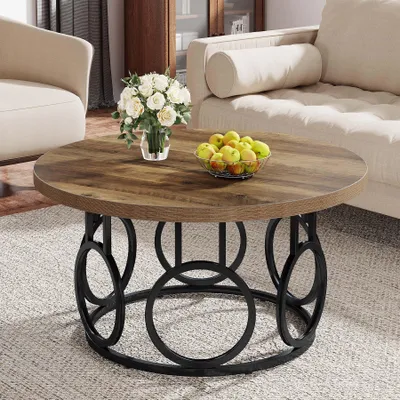 A round coffee table with a wood grain top and a black metal base, decorated with a vase of white flowers and a bowl of apples, placed on a rug in a living room setting.