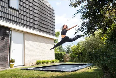 A young woman is performing a split jump in the air above a rectangular trampoline in a backyard setting.