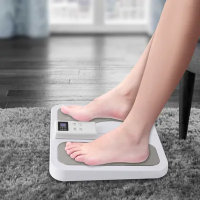 A person's legs and feet are shown on a white and gray foot massager placed on a gray rug.