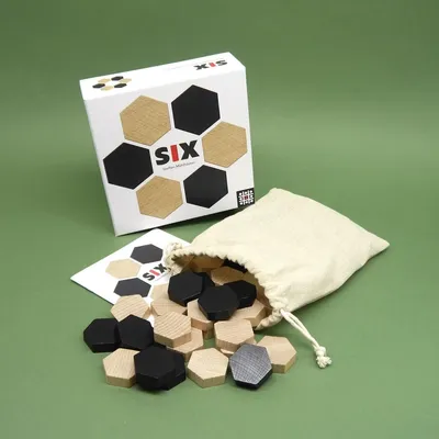 Schachtel und Beutel des taktikspiel „SIX“; zahlreiche schwarze und naturfarbene Hexagon-Steine sind ausgestreut.