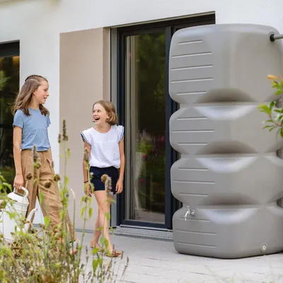Two young girls are standing on a patio next to a large gray water tank.